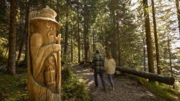 Twee wandelaars bekijken gebeeldhouwde houten figuren op het "pad van de Walser legendes", een themapad midden in het Alpenlandschap met uitzicht op de bergen