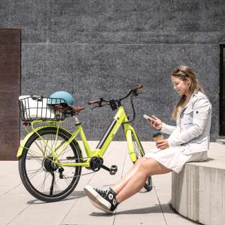 Jonge vrouw zit met koffie en smartphone naast een gele e-bike in het stadscentrum van Vaduz