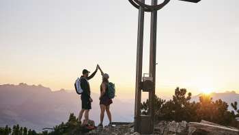 Twee wandelaars geven elkaar een high-five bij het topkruis op de Alpspitz bij zonsopgang met een bergpanorama op de achtergrond.