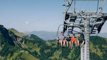 Een gezin rijdt met de Sareis-stoeltjeslift over een groen berglandschap