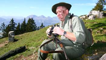 Een jager met een verrekijker zit in een weiland tegen de achtergrond van een alpine berg.