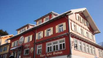 Buitenaanzicht van het Alpenhotel Vögeli Malbun met de Elchbar op de begane grond - traditioneel rood gebouw met witte letters tegen een blauwe lucht.