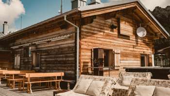 Gezellig zonneterras met houten banken en comfortabel loungemeubilair voor de Sareis berghut op 2.000 meter hoogte, met stralende zon en een bergachtige achtergrond.