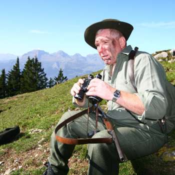 Een jager met een verrekijker zit in een weiland tegen de achtergrond van een alpine berg.