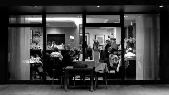 Zwart-witfoto van een druk restaurant met uitzicht van buitenaf door de glazen pui.