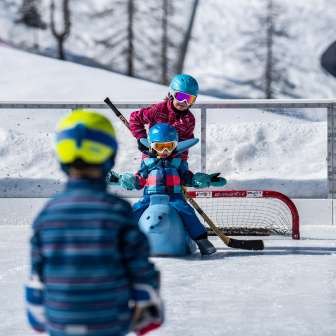 Kinderen spelen met helmen en hockeysticks op de ijsbaan in Malbun, een kind zit op een schaatshelper voor het doel.