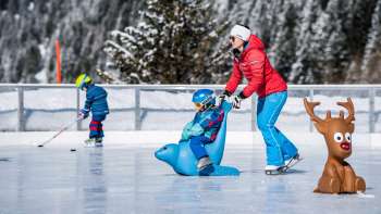 Familie op de ijsbaan bij Schlucher-Treff Malbun, ondersteund door een schaatshulp in de vorm van een blauw dier.