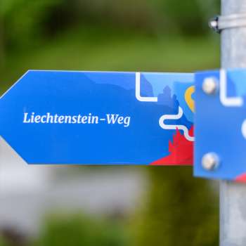 Blauwe wegwijzer met het opschrift "Liechtenstein-Weg" in een modern ontwerp.