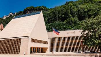 Modern parlementsgebouw in Vaduz met een puntgevel, de Liechtensteinse vlag ervoor en omgeven door groene heuvels. Modern parlementsgebouw in Vaduz met een puntgevel, de Liechtensteinse vlag ervoor en omgeven door groene heuvels.