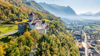 Luchtfoto van het kasteel van Vaduz op een heuvel boven de stad, met op een heldere dag een panoramisch uitzicht op de Alpen en de Rijnvallei.