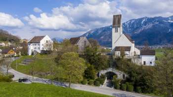 Panoramisch uitzicht op de "Kirchhügel" (kerkheuvel) van Bendern met zijn historische kerk, omgeven door goed onderhouden paden, bomen en traditionele architectuur tegen een alpiene bergachtergrond.