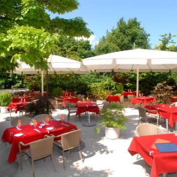 Buitenaanzicht van het terras met rode tafelkleden, omringd door bomen en parasols.