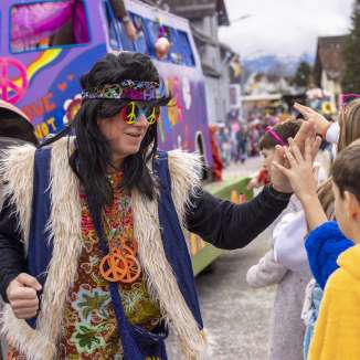 Hippie verkleed op het kindercarnaval in Mauren, high-fives met kinderen voor een kleurrijke praalwagen.
