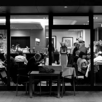 Zwart-witfoto van een druk restaurant met uitzicht van buitenaf door de glazen pui.
