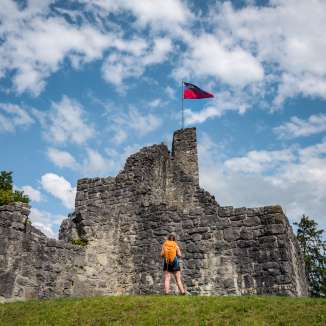 Wandelaar voor de historische ruïne van kasteel Schellenberg met de Liechtensteinse vlag onder een blauwe hemel