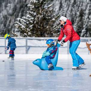 Moeder duwt kind op een schaatsende pinguïn over de ijsbaan in Malbun, op de achtergrond een kind met een ijshockeystick