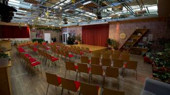 Grote, met licht overgoten evenementenruimte van de Kreativ Akademie met podium, rode gordijnen en rijen stoelen - ideaal voor seminars, lezingen en creatieve evenementen in Liechtenstein.