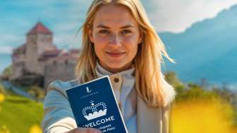Vrouw houdt de "WELCOME Belevenispas" omhoog voor Kasteel Vaduz in een bloeiend veld
