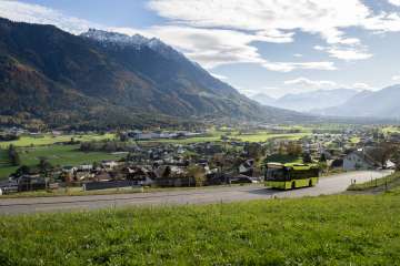 Groene LIEmobil-bus rijdt over een kronkelige weg boven Liechtenstein met uitzicht op het Rijndal en de omringende Alpen