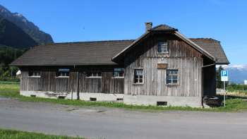 Historisch houten gebouw van de oude zagerij in Schaanwald tegen een alpine achtergrond onder een blauwe hemel.