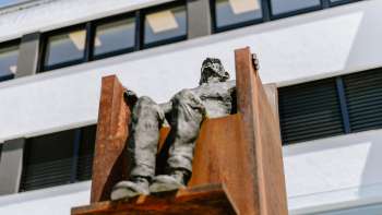 Sculptuur "Hochsitz" van Robert Indermaur: figuur zit verheven op een roestkleurige stalen stoel voor een moderne gevel.
