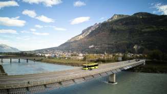 Gele LIEmobil-bus rijdt over de Rijnbrug bij Vaduz met een bergpanorama op de achtergrond - Beleef Liechtenstein