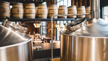 Zicht op een moderne brouwerij met glimmende roestvrijstalen tanks en gestapelde houten vaten op de achtergrond.