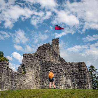 Wandelaar voor de historische ruïne van kasteel Schellenberg met de Liechtensteinse vlag onder een blauwe hemel