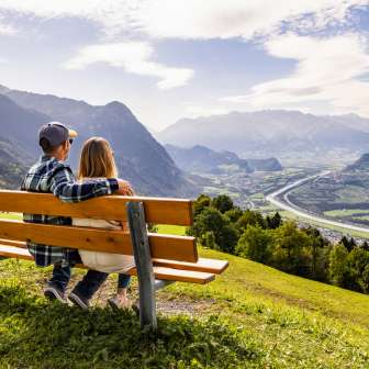 Echtpaar zittend op een bankje in Triesenberg en uitkijkend over de Rijnvallei in Liechtenstein met een indrukwekkend Alpenpanorama