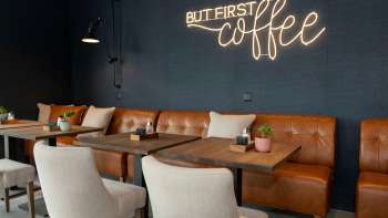Een modern café met leren banken, witte fauteuils en houten tafels. Het opschrift "BUT FIRST coffee" schittert in felle neonletters op de donkerblauwe muur. Een modern café met leren banken, witte fauteuils en houten tafels. Het opschrift "BUT FIRST coffee" schittert in felle neonletters op de donkerblauwe muur.