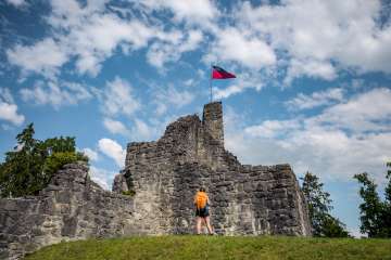 Wandelaar voor de historische ruïne van kasteel Schellenberg met de Liechtensteinse vlag onder een blauwe hemel