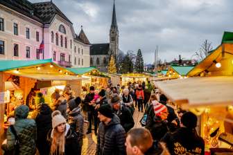 Sfeervolle avondopname van de kerstmarkt in Vaduz 2018 met feestelijk versierde marktkramen en mensenmassa's voor het regeringsgebouw en de kathedraal