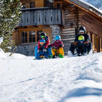 Twee volwassenen en drie kinderen zitten op sleeën en beginnen samen aan een afdaling voor een besneeuwde berghut in Malbun